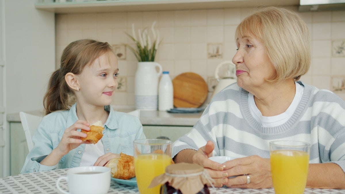 Kid adorable girl is eating croissant and talking to happy granny laughing having breakfast at kitchen table. Family relationship and childhood concept.
